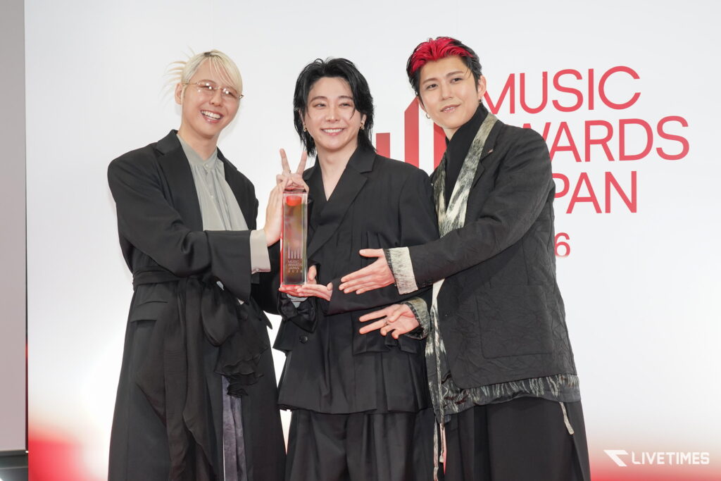 Three award winners posing together, holding a tall glass trophy and smiling at a music awards event against a white backdrop with red text partially reading 'MUSIC AWARDS JAPAN'.