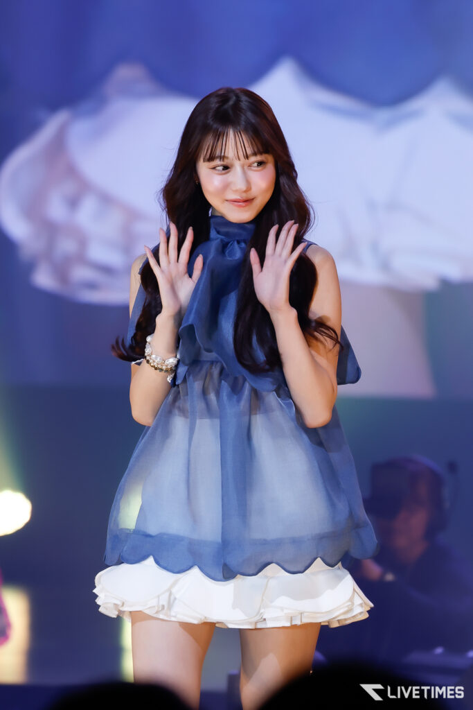 Female performer on stage, wearing a blue sleeveless dress, smiling and waving with both hands raised.