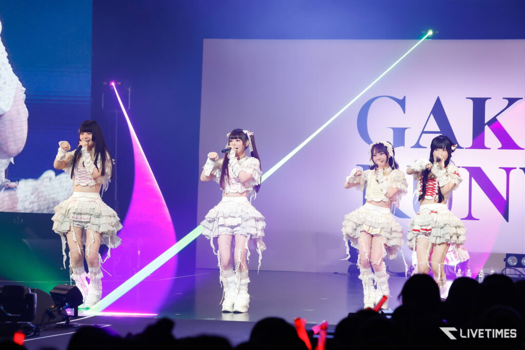 Four female pop idols perform on a stage in frilly white outfits with microphones, colorful neon lights in the background and an audience in the foreground.