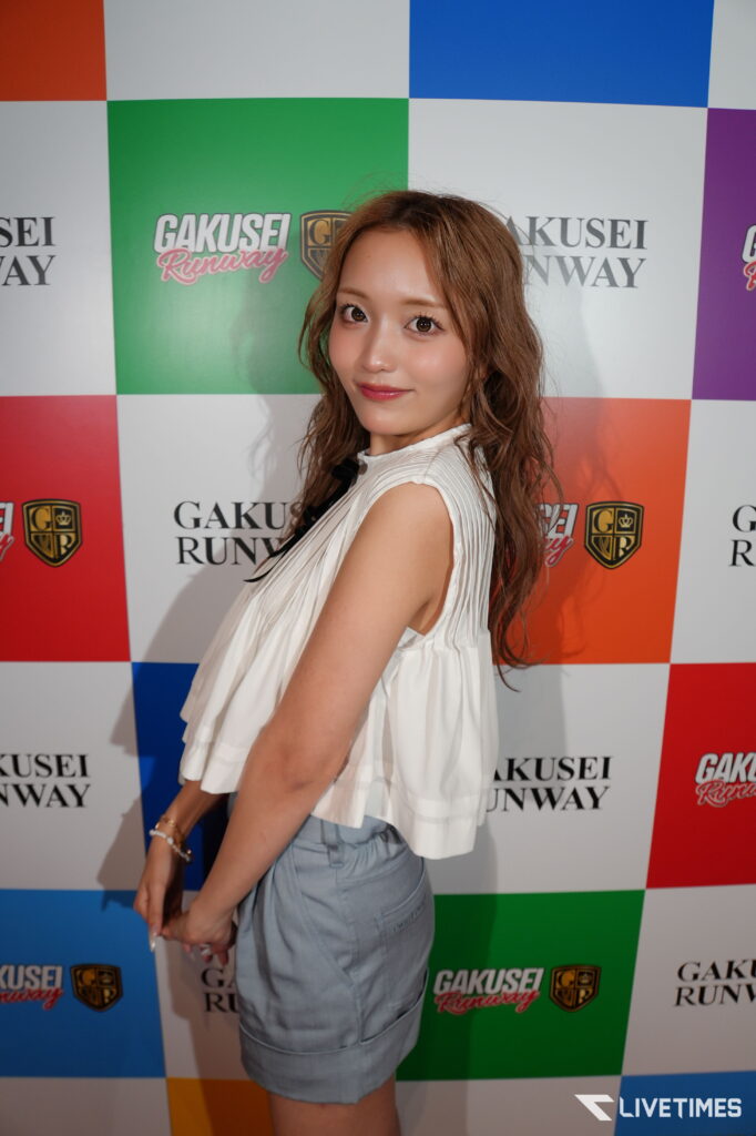 Young woman posing at a Gakusei Runway event, facing camera with a white pleated top and light blue skirt, colorful backdrop behind her