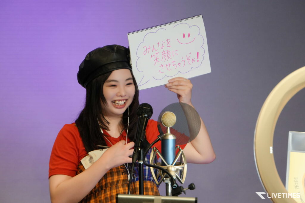 Smiling person on stage holds a white sign with pink Japanese text and a cloud outline.