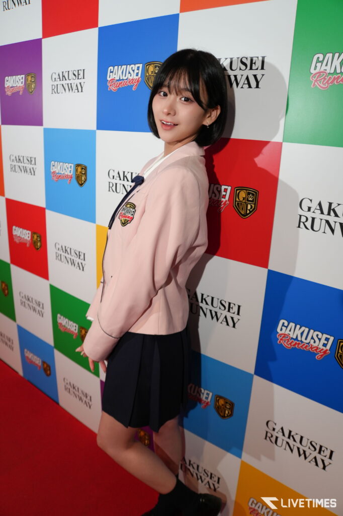 Young woman with short black hair, wearing a pale pink blazer and navy skirt, posing on a multicolor checkerboard backdrop that says GAKUSEI RUNWAY.