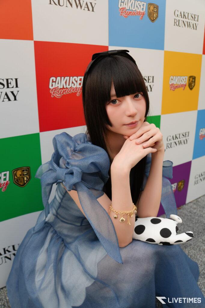 Young woman with black hair and a bow sits with hands clasped under her chin in front of a colorful checkered backdrop that reads 'GAKUSEI RUNWAY'.