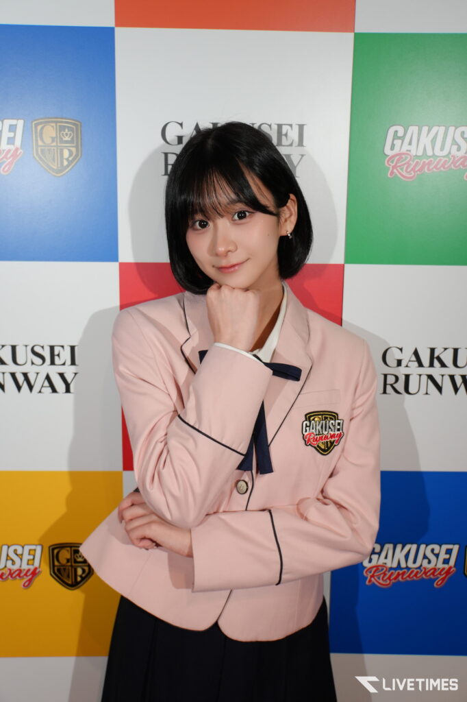 Young woman in a pale pink blazer with a navy bow, resting her chin on her hand at a colorful backdrop that reads GAKUSEI RUNWAY.