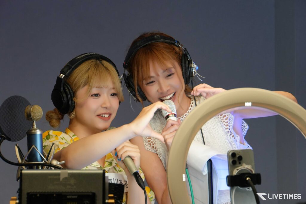 Two women wearing large over-ear headphones share a microphone in a recording studio, smiling and pointing toward the equipment as they sing or speak.