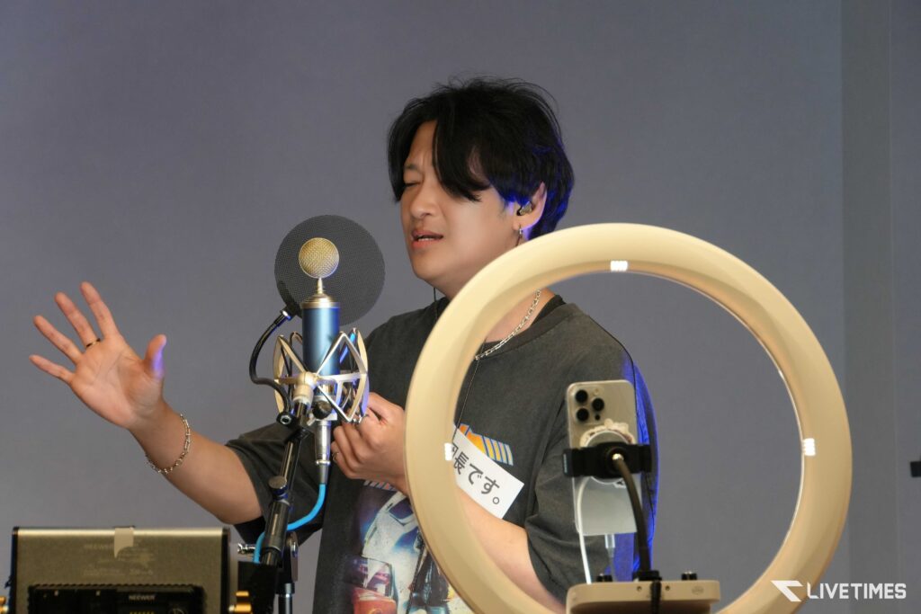 Person singing into a studio microphone with a pop filter, ring light in the foreground, and a hand raised as if gesturing to emphasize the performance.