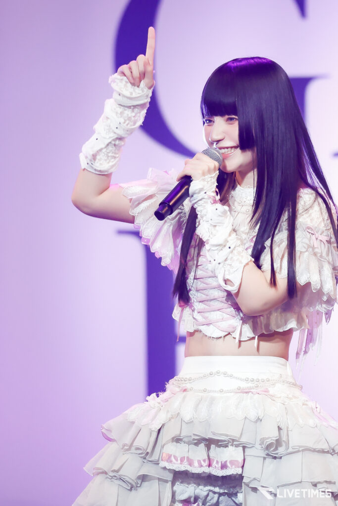 Female pop singer on stage, holding a microphone and raising one finger, dressed in a pink and white frilly outfit.