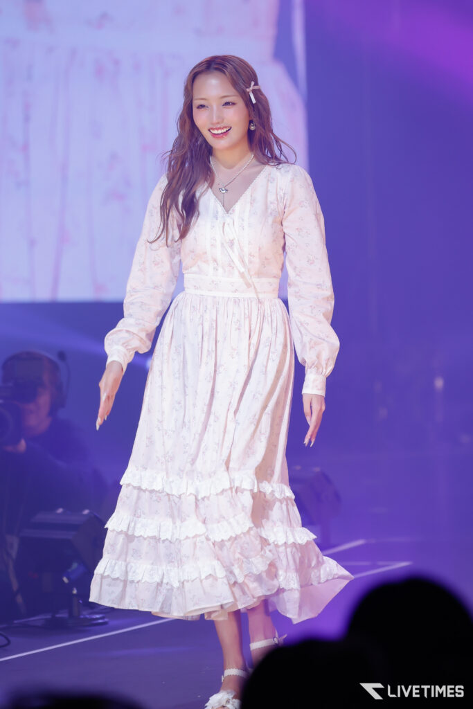 Smiling female performer on stage wearing a pale pink floral dress with ruffled layers and a hair clip, under pink-purple lighting.