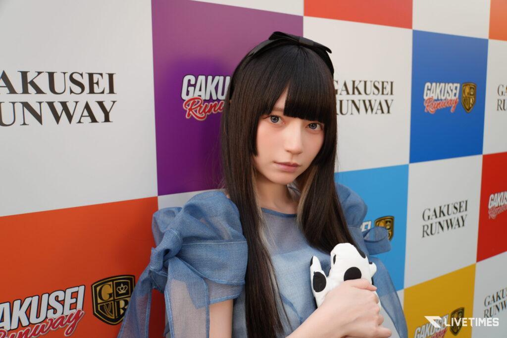 Woman with long dark hair in a blue outfit posing in front of a colorful GAKUSEI RUNWAY backdrop, holding a panda plushie.
