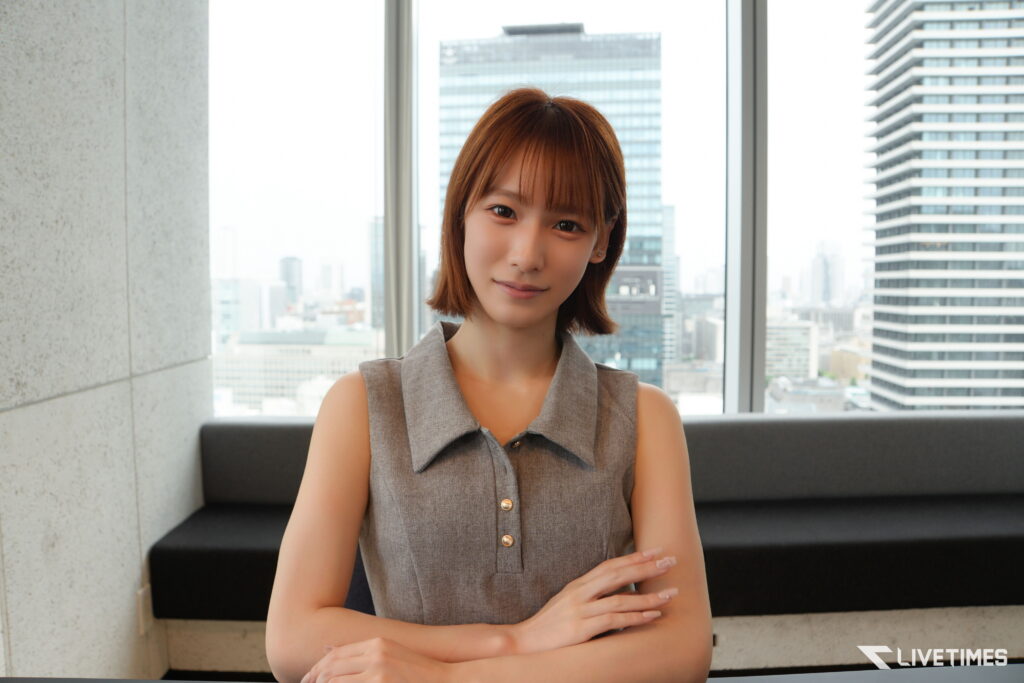 Woman in a gray sleeveless blouse sits on a couch in a modern office with a city skyline outside the window.