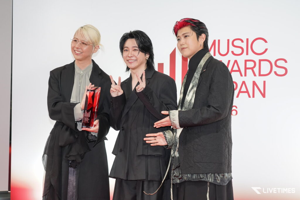 Three people in black outfits pose together at a Music Awards Japan event, with one holding a glass trophy and flashing peace signs.