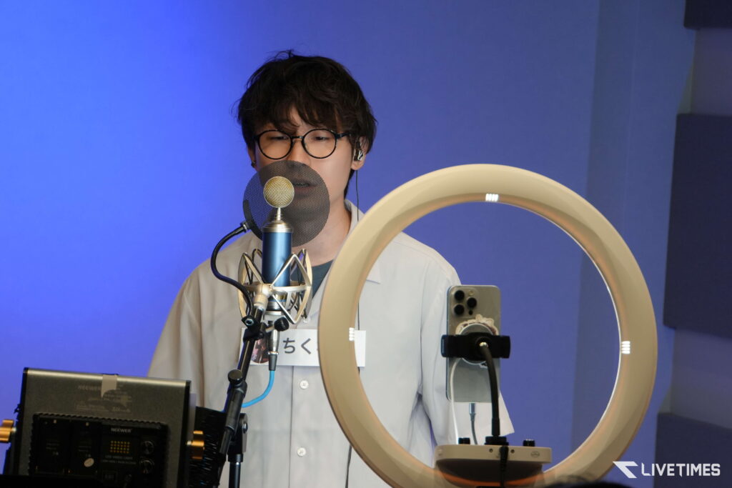 Person in glasses speaks into a large microphone with a pop filter in a blue studio background (recording setup).