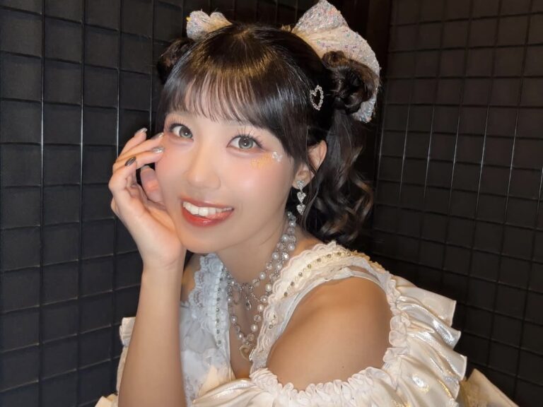 Close-up portrait of a young woman smiling, with updo buns, glitter makeup, and a white ruffled dress accessorized with pearls and bows.