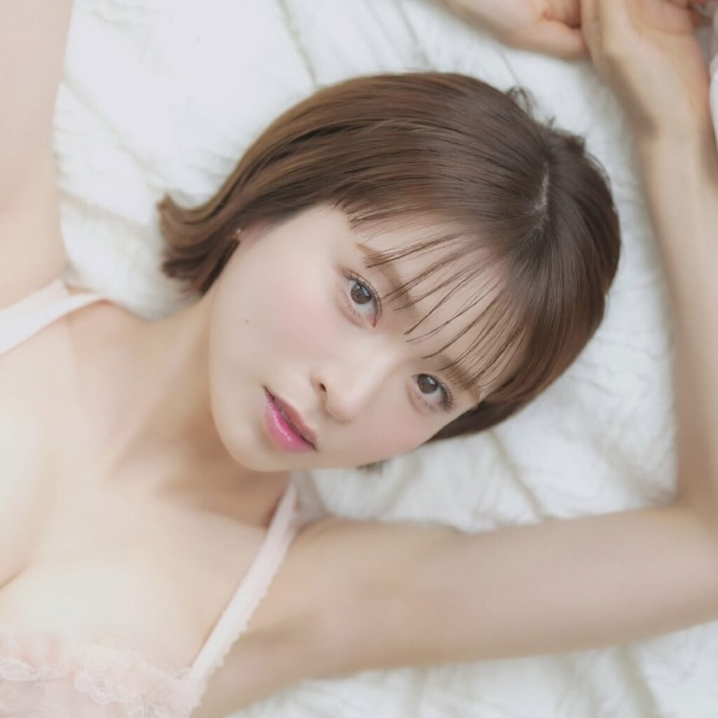 Close-up of a young woman with brown hair lying on a white bed, facing the camera in a pink lace camisole.