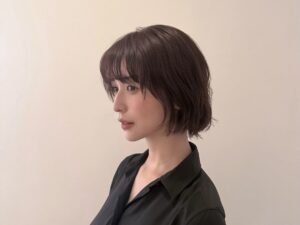 Side profile of a young woman with a short, dark brown bob haircut wearing a black shirt against a beige wall