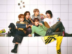Five young friends sitting on a white tiled wall, snacking and sharing drinks in a casual setting with posters taped on the wall behind them.