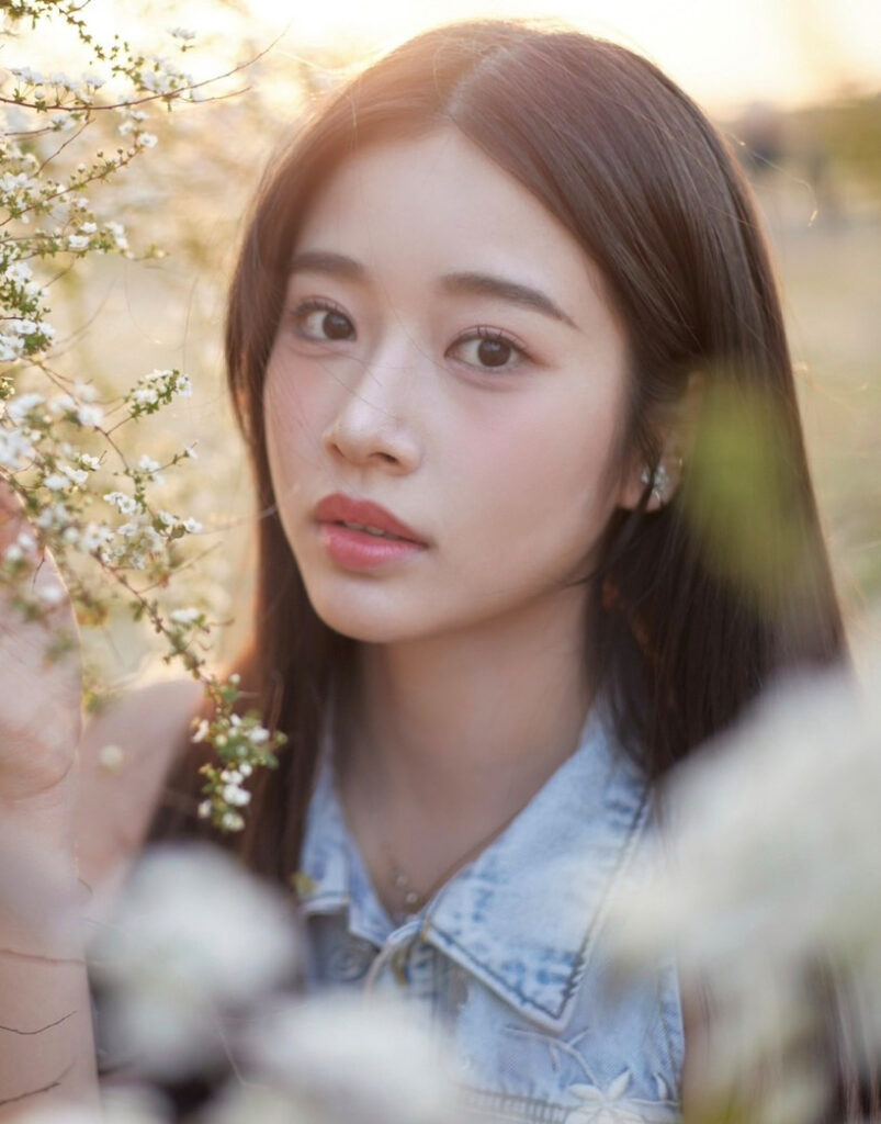 Close-up portrait of a young woman with long dark hair among white blossoms in warm sunlight.