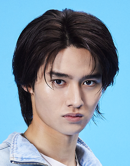 Close-up portrait of a young man with dark hair and a denim jacket, looking straight at the camera against a bright blue background.