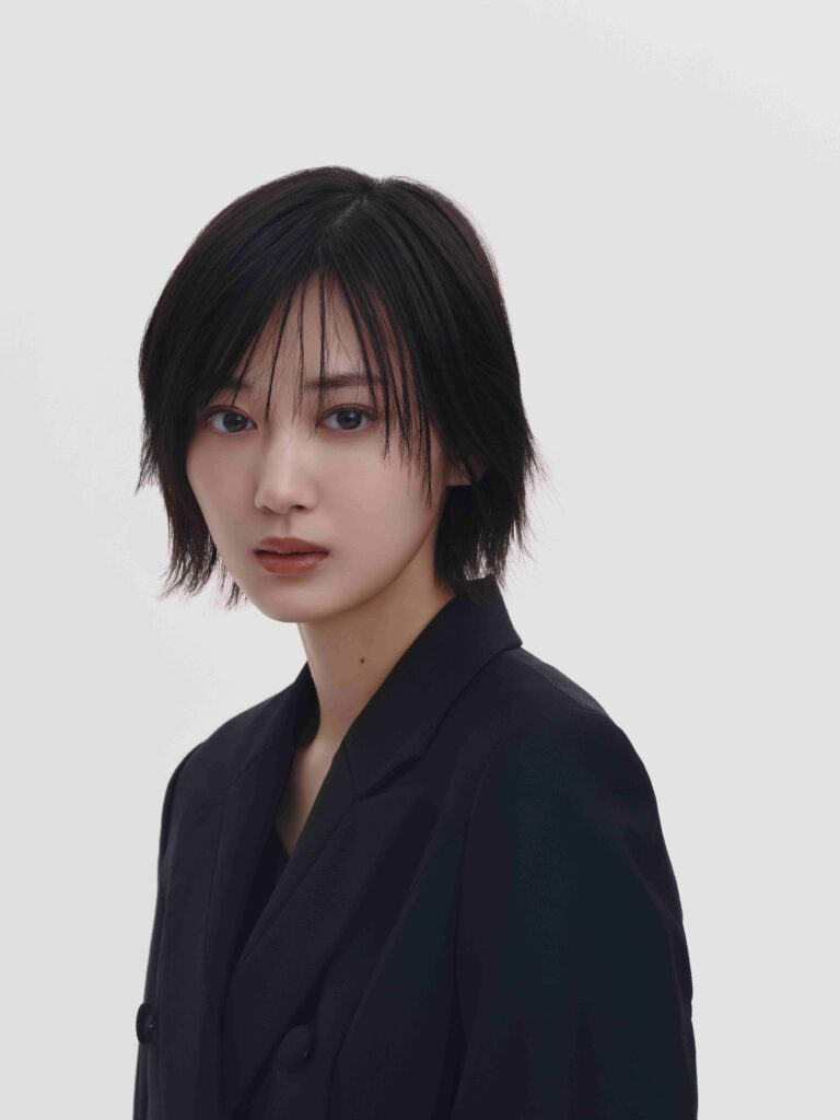 Portrait of a person with short black hair wearing a dark blazer against a light gray background, looking at the camera at a slight angle.