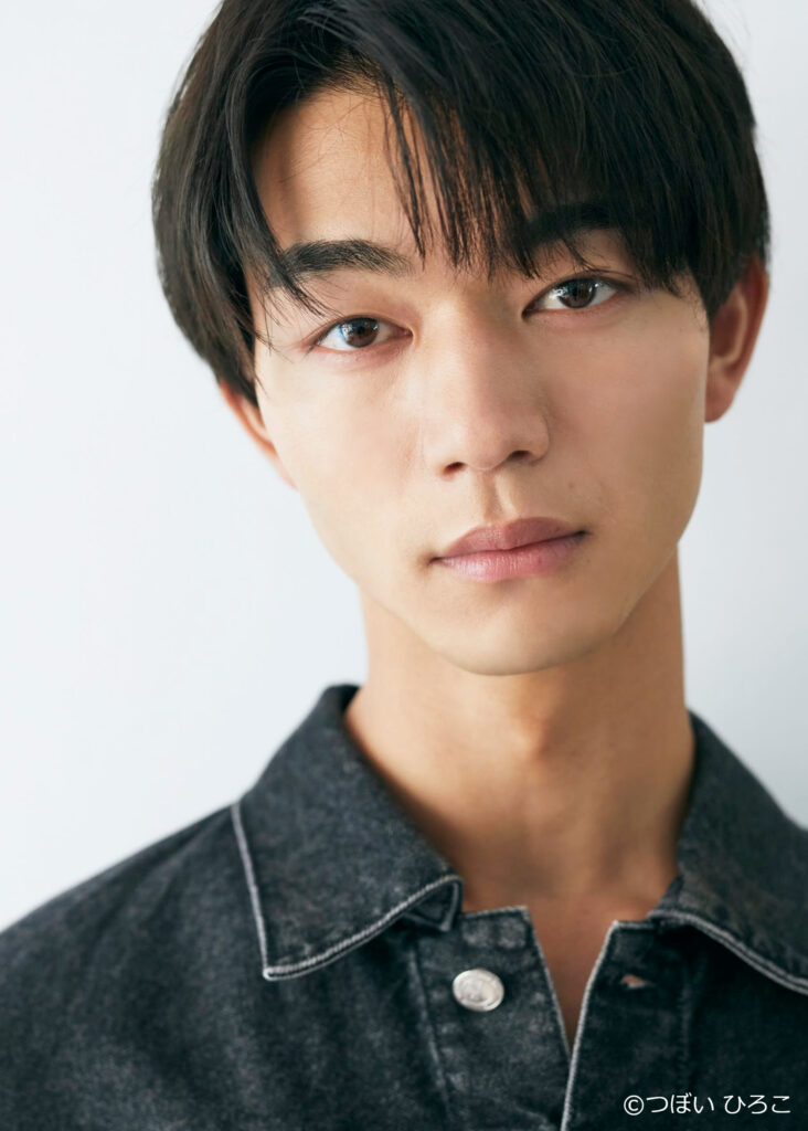 Close-up portrait of a young man with dark hair and brown eyes, wearing a dark denim shirt; subtle smile, neutral expression.