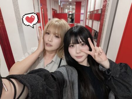 Two women take a selfie in a red-and-white hallway; one with blonde hair touches her cheek while the other with black hair flashes a peace sign nearby.
