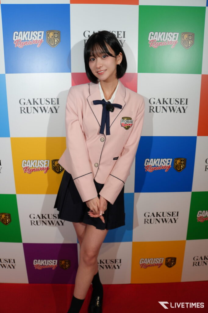 Young woman in a pale pink blazer and navy bow posing at a colorful Gakusei Runway backdrop with the event logo behind her.