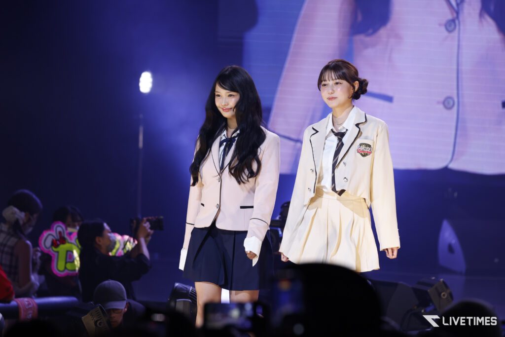 Two young performers in matching cream school uniforms walk on stage under blue lights while fans in the front row take photos.