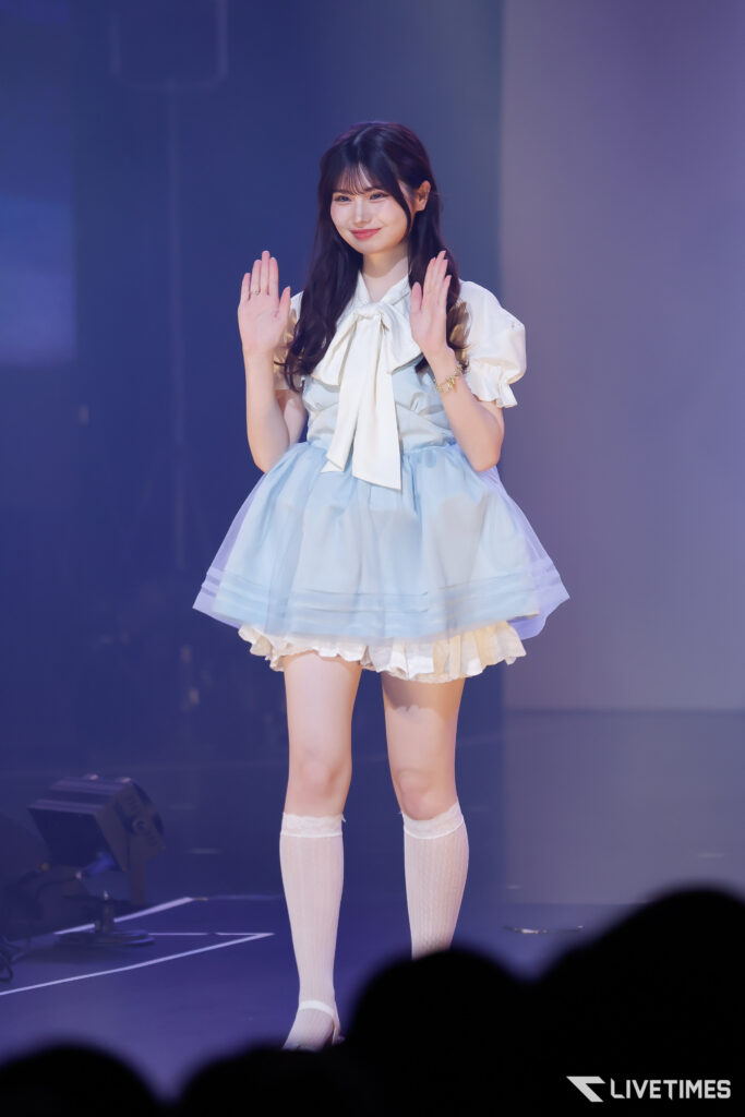 Young woman in a light blue ballroom-style dress with a large white bow waves to the audience onstage, smiling warmly.