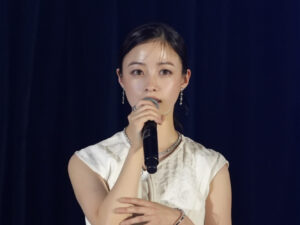 Young woman in a white sleeveless blouse singing into a microphone on stage against a dark blue backdrop, wearing jewelry.