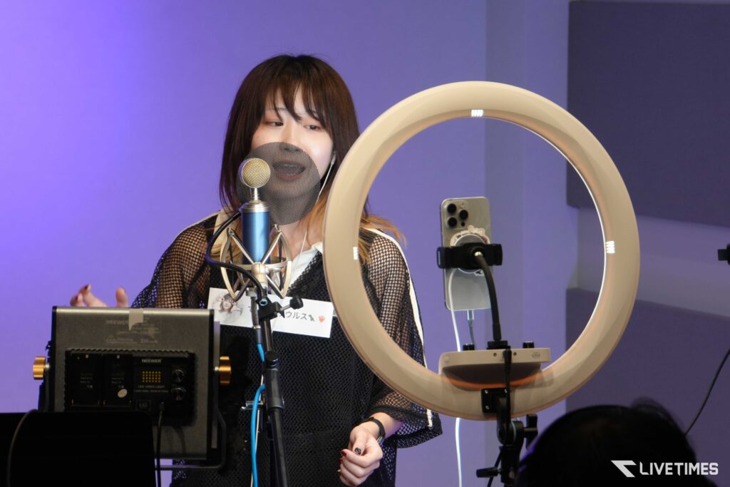 Woman recording vocals into a blue microphone with a mesh pop filter in a studio with a circular ring light nearby.