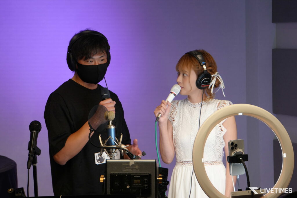 Two people in a recording studio singing into microphones, wearing headphones; purple backdrop behind them.