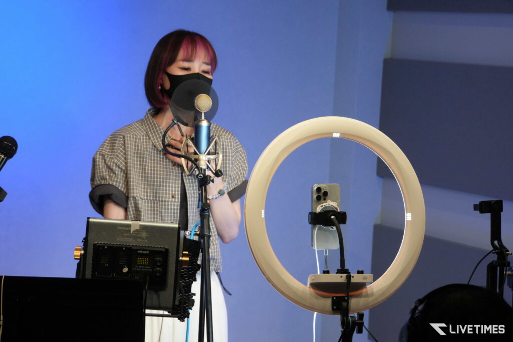 Person wearing a black face mask singing into a studio microphone with a pop filter, surrounded by recording equipment and a circular ring light in a blue room.