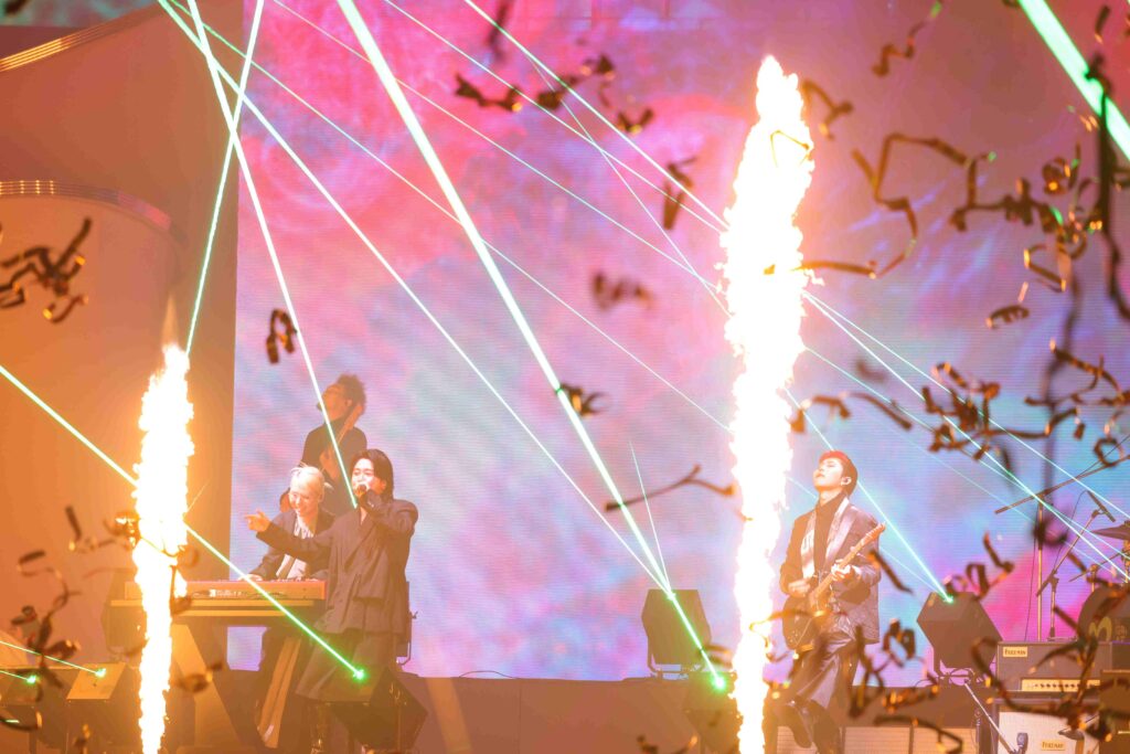 Live concert scene with musicians on stage, green lasers crossing the air and flames shooting upward amid colorful lights and confetti in the foreground.