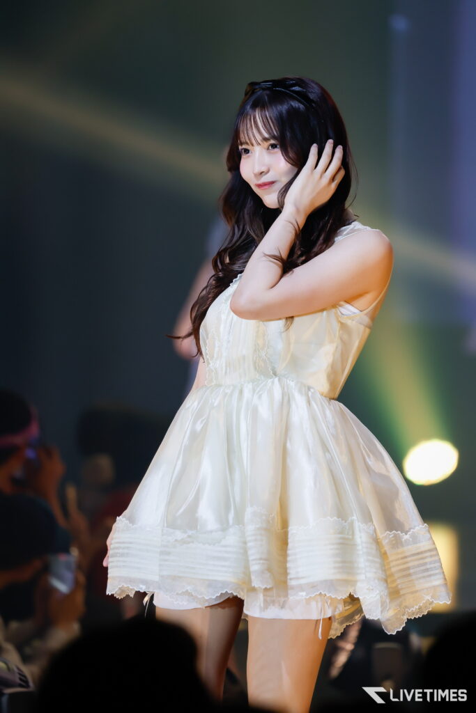 Young woman in a white dress on stage, touching her hair and looking slightly to the side during a performance.