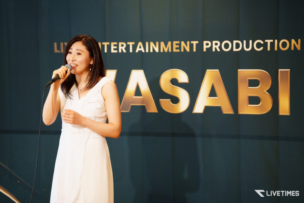 Female singer in a white dress singing into a microphone on a stage with a dark blue backdrop and large gold letters behind her (LIVETIMES watermark).