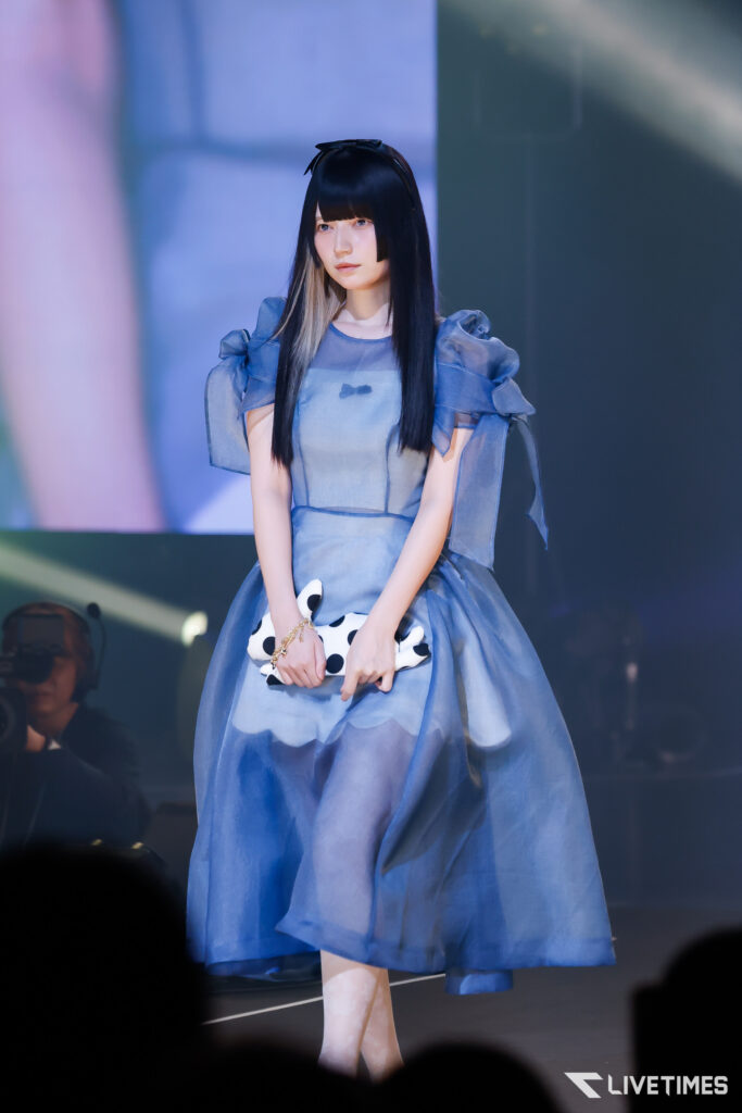 Female performer in a blue, puff-shoulder dress on stage, holding a white polka-dot clutch with a blue bow detail.