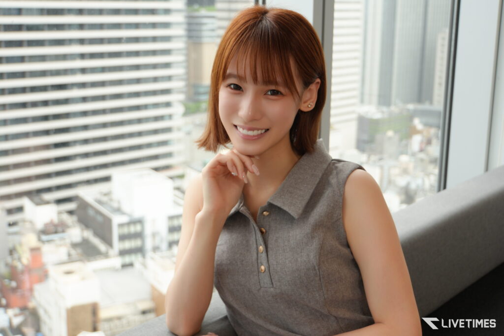 Smiling woman with short brown hair sits by a window overlooking a cityscape in a high-rise building.