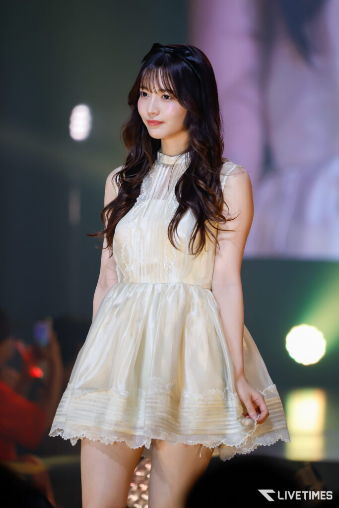 Female performer with long dark hair in a cream lace dress on stage, soft smile, colorful lights in the background.