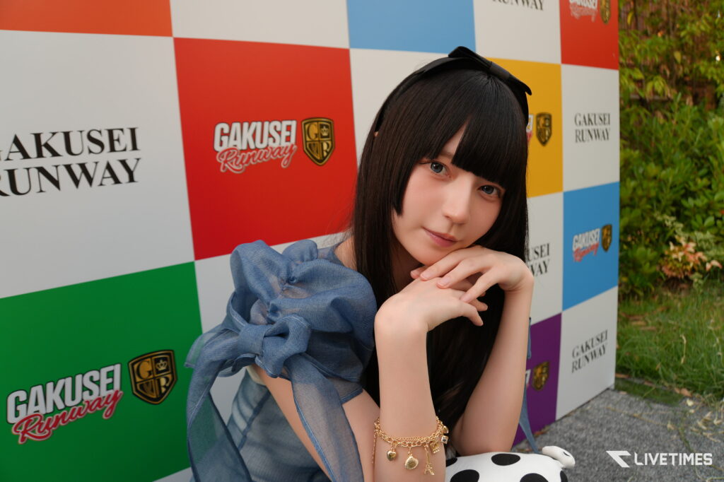 Portrait of a young woman with long black hair posing with her chin resting on her hands against a colorful checkered wall that reads 'Gakusei Runway'.