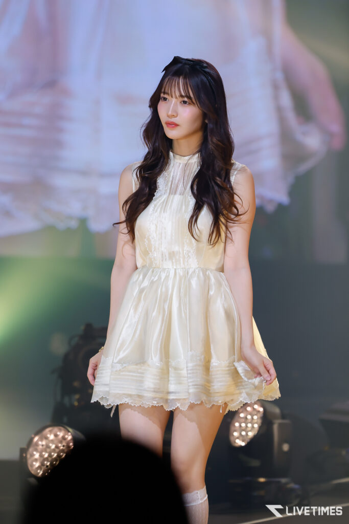 Young woman in a cream lace dress standing on stage under soft lights during a performance.