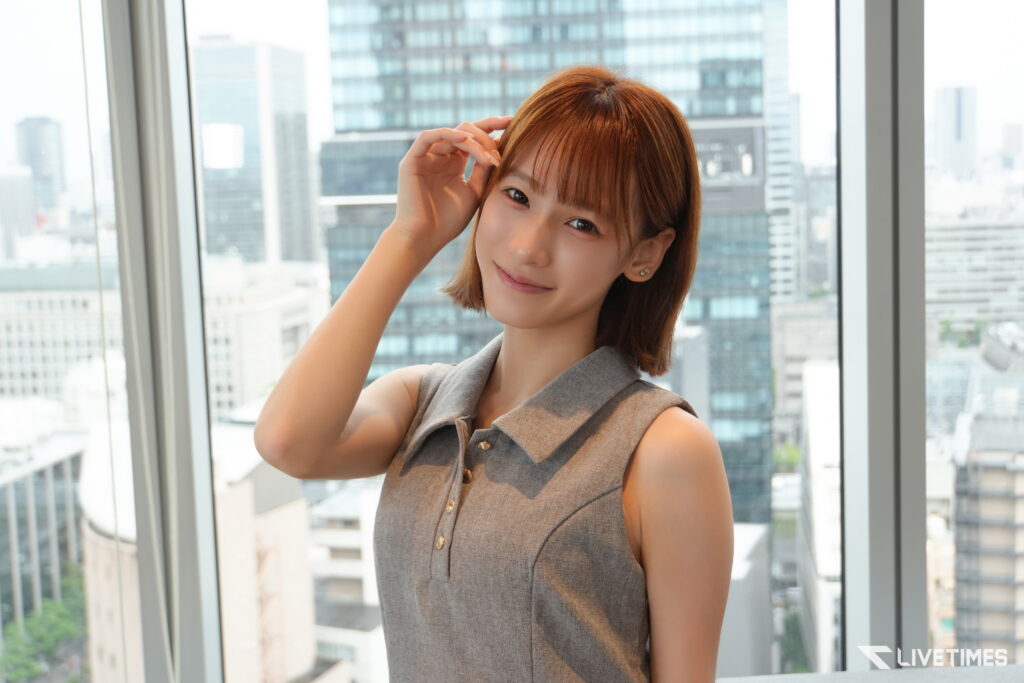 Woman smiling and posing near large windows with a city skyline in the background, wearing a gray sleeveless blouse/dress.