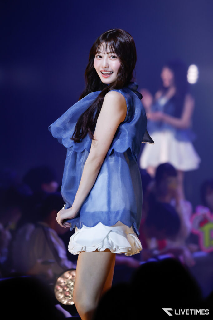 A female performer in a blue sleeveless top and white ruffled skirt on stage, smiling at the audience.