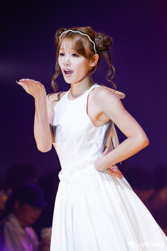 Singer in a white sleeveless dress performing on stage, with a pearl headband and styled buns, hand raised mid-performance.