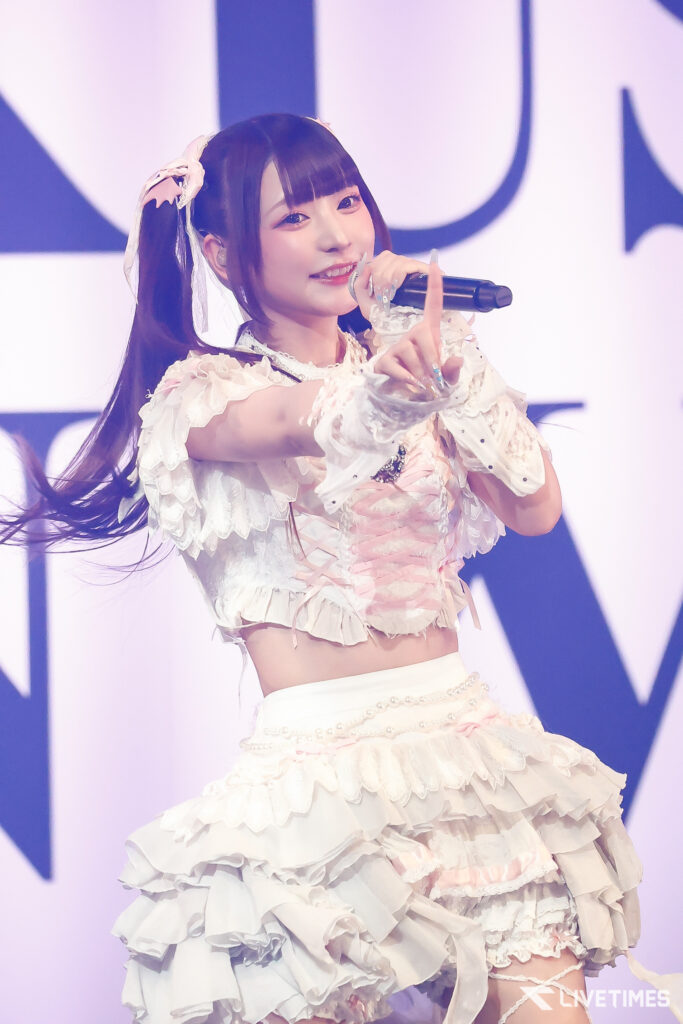 Young female performer on stage wearing a white frilly dress with ribbons, holding a microphone and pointing to the audience