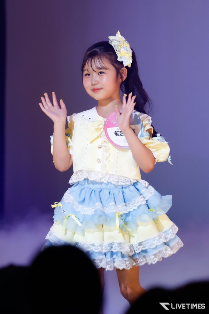 Young girl in a pastel yellow and blue frilly dress waves on stage, wearing a decorative hair bow and a round badge on her chest.