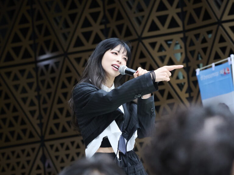 Female singer in a pinstripe blazer sings into a microphone while pointing toward the audience, stage backdrop behind her.