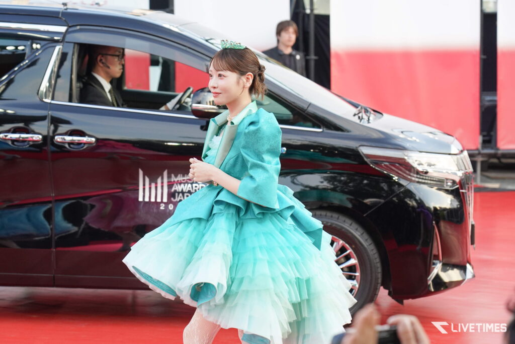 Woman in a teal-green ball gown with a tiara walks along a red carpet beside a black SUV at a formal event.