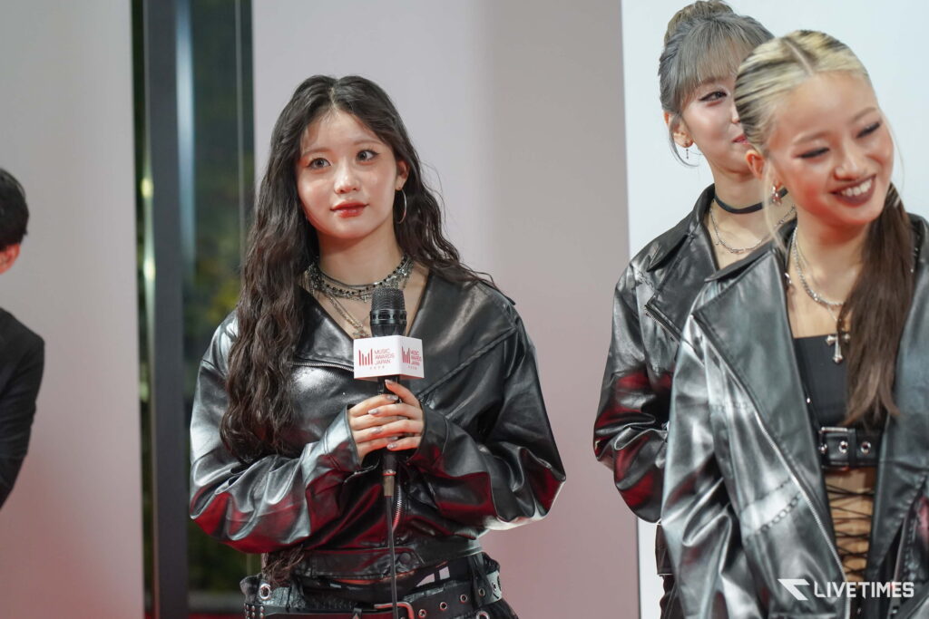 Woman in a shiny black leather outfit holding a microphone at a press event with others smiling nearby.