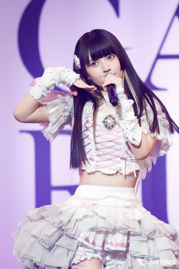 Singer on stage with a microphone, wearing a frilly pink-and-white dress under purple lighting, performing live.