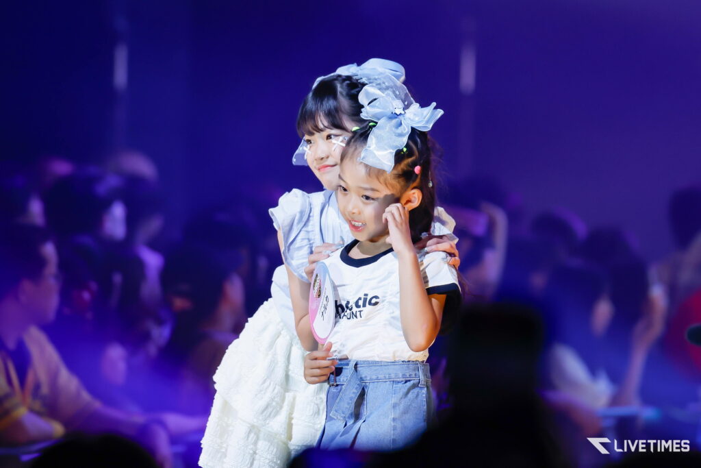Two young girls on stage—one piggybacks the other—smiling, with matching light blue bows and white outfits, a blurred audience in purple lighting behind them.